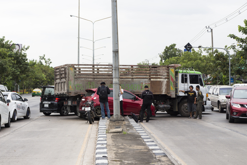 Several vehicles involved in a traffic accident at an intersection, including a red car and a large truck, with people inspecting the scene on a divided road.