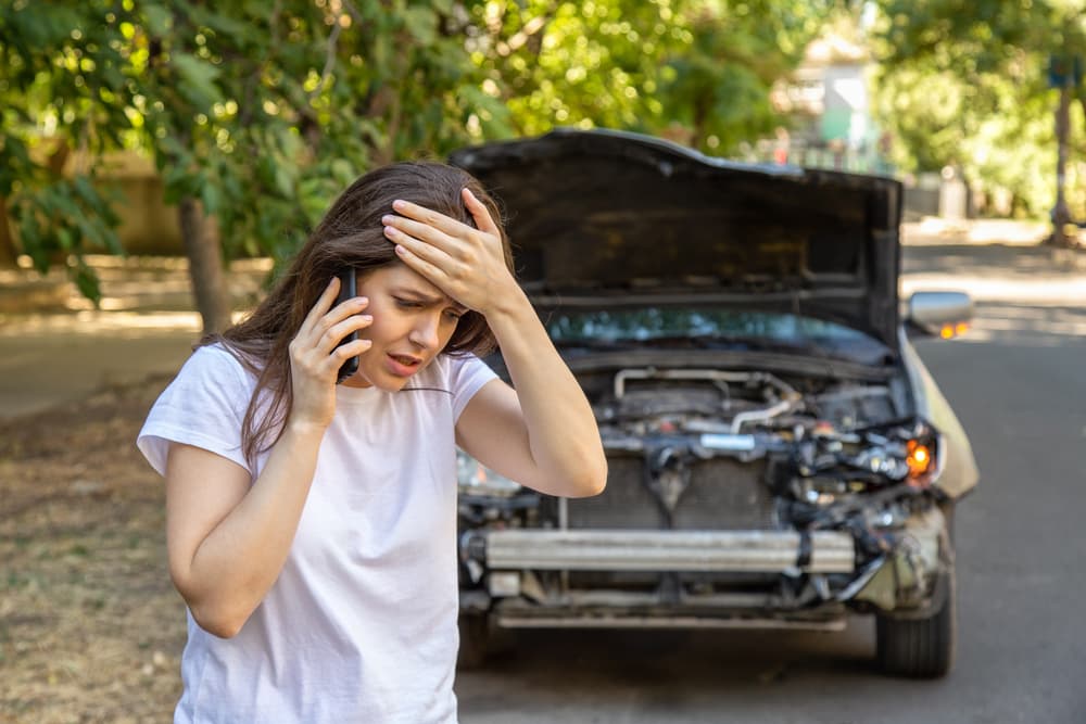 Woman driver in distress by wrecked car after accident, seeking help from auto insurance. Stressful aftermath of a car crash.