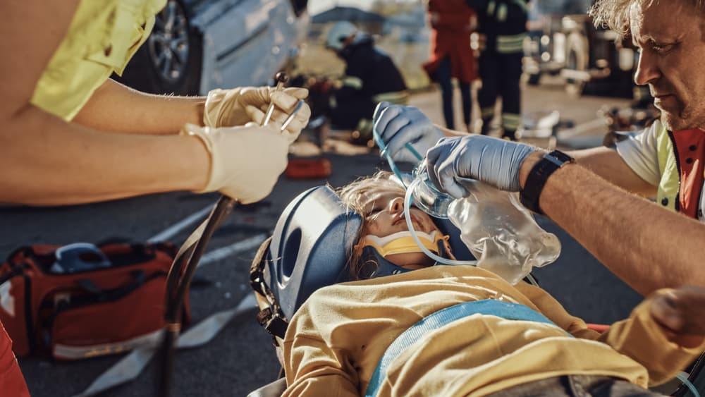 Paramedics attending to a car crash victim on stretchers, administering first aid and oxygen on the accident scene.