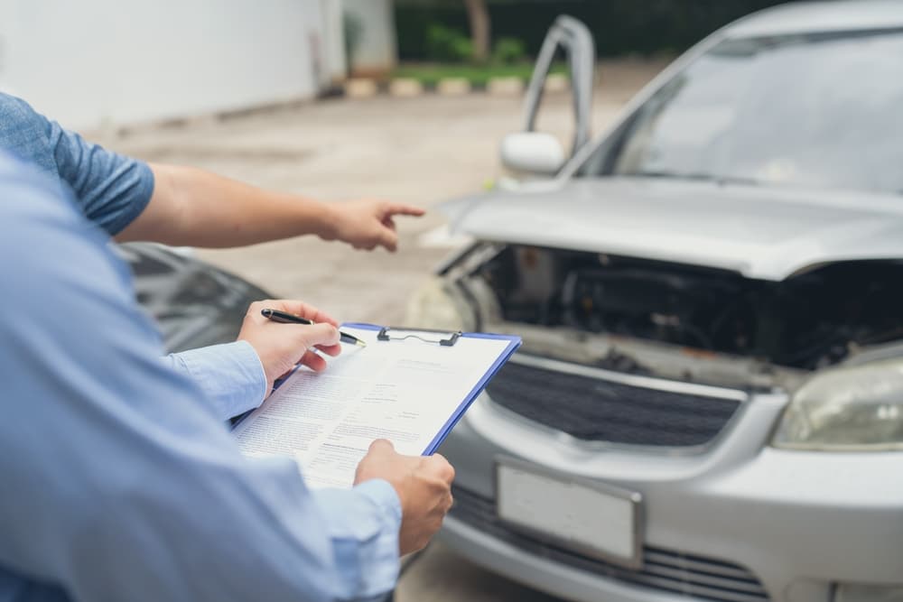 Insurance officer writing a claim report on clipboard during a car crash insurance process with a man and an insurance agent.