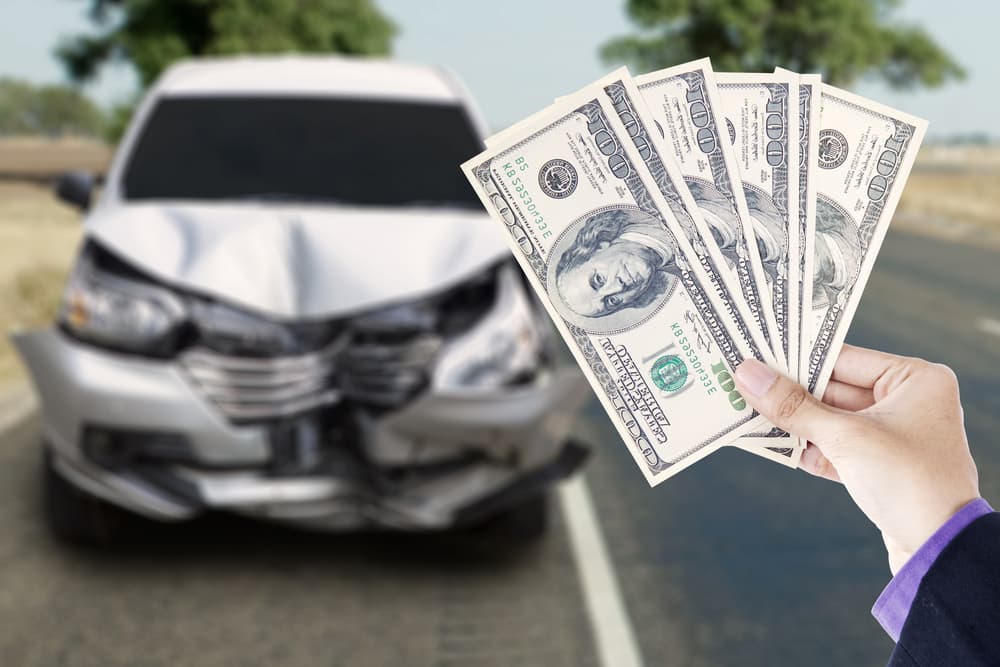 Businessman's hands holding dollar bills with a broken car on the highway - financial loss after a car accident