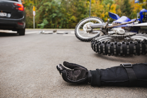 A close-up of a motorcycle lying on the ground after an accident, with the rider's gloved hand outstretched and a car parked nearby, indicating a crash scene.