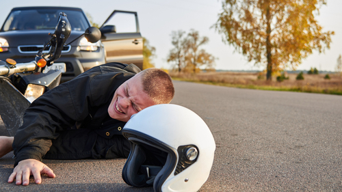 A motorcyclist lying injured on the road near his fallen motorcycle, with a white helmet nearby and a car stopped with its door open in the background.