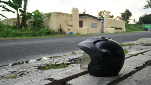 A black motorcycle helmet placed on the side of a road, with a rural house and greenery in the background, symbolizing a potential motorcycle accident scene.