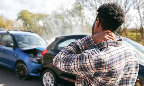 A man in a plaid shirt is standing next to a car accident scene, holding his neck in pain, possibly suffering from whiplash after a rear-end collision.