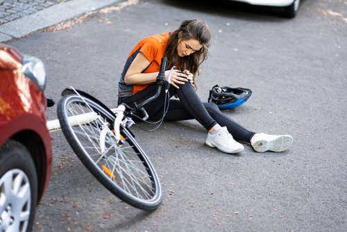 A young woman sits on the ground next to her bicycle, holding her injured arm after being hit by a car, with her helmet lying beside her on the street.