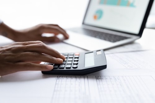 A close-up of a person using a calculator while working on financial documents with a laptop open in the background.