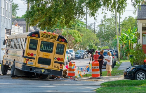 A yellow school bus partially off the road, having crashed into construction barriers, while onlookers, including a news crew, observe the scene.