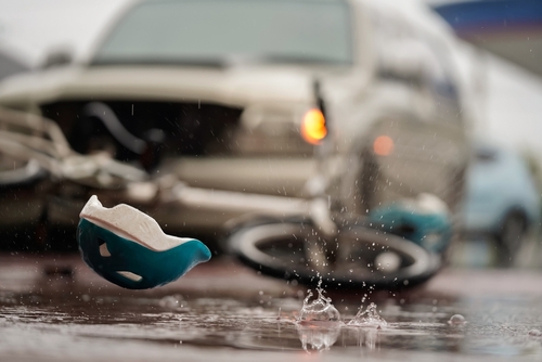 A close-up shot of a bicycle helmet lying in a puddle on the road, with a damaged car in the background, highlighting a recent accident in rainy conditions.