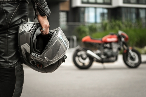 A person dressed in a leather jacket holding a motorcycle helmet, with a motorcycle parked on a street in the background, indicating readiness for a ride.