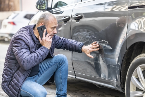 An older man inspecting a scratch on the side of his car while speaking on his smartphone, likely discussing the damage with his insurance company.