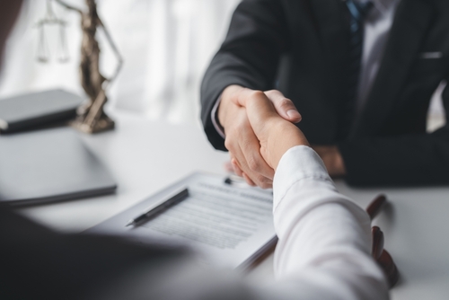 A close-up of a handshake between two individuals in a professional setting, with legal documents on a desk in the background, symbolizing agreement or partnership.