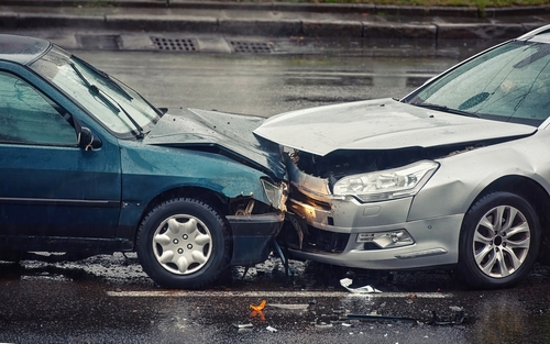 Two cars have collided head-on in an accident on a wet road, with visible damage to the front ends of both vehicles.