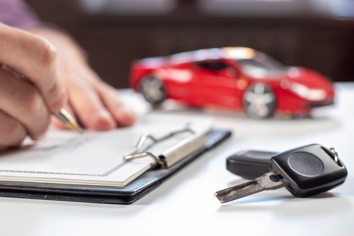 A close-up of a person signing a document on a clipboard with a red toy car and car keys in the background, symbolizing car insurance or car purchase agreements.