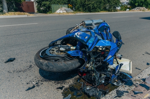 A heavily damaged blue motorcycle lying on its side on the road after a severe accident, with debris scattered around, indicating the aftermath of a collision.