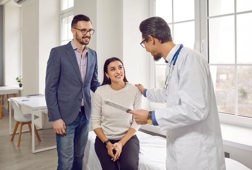 A doctor is discussing medical results with a smiling female patient, who is seated on an examination table, while her partner stands beside her in a supportive manner.