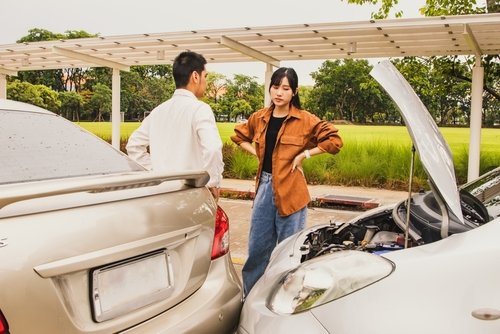 A woman and a man stand beside two cars parked under a shelter, discussing a mechanical issue with one of the cars. The woman, dressed in a casual brown jacket and jeans, appears to be explaining or questioning something about the car’s engine.