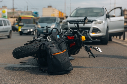 A motorcycle lying on the ground after a traffic accident, with a car in the background and a black backpack near the fallen motorcycle.