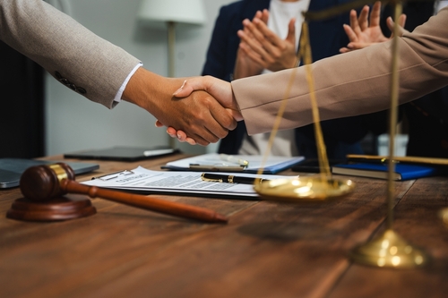 A professional setting where two individuals shake hands across a table in an agreement or settlement, with others clapping in the background, suggesting a successful negotiation. A gavel and legal documents are visible, emphasizing the legal or formal nature of the event.