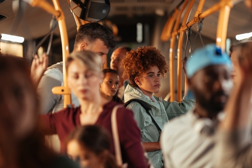 A crowded bus with passengers standing and holding onto handrails, with a focus on a young woman with curly hair looking out of the window.