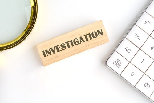 A wooden block labeled "INVESTIGATION" next to a magnifying glass and a calculator on a white surface, symbolizing the process of analyzing or examining details.