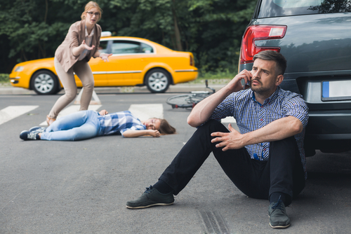 A car accident scene where a man sits on the curb, visibly in shock, using his phone, while a woman lies injured on the road and another person attends to her, with a taxi in the background.