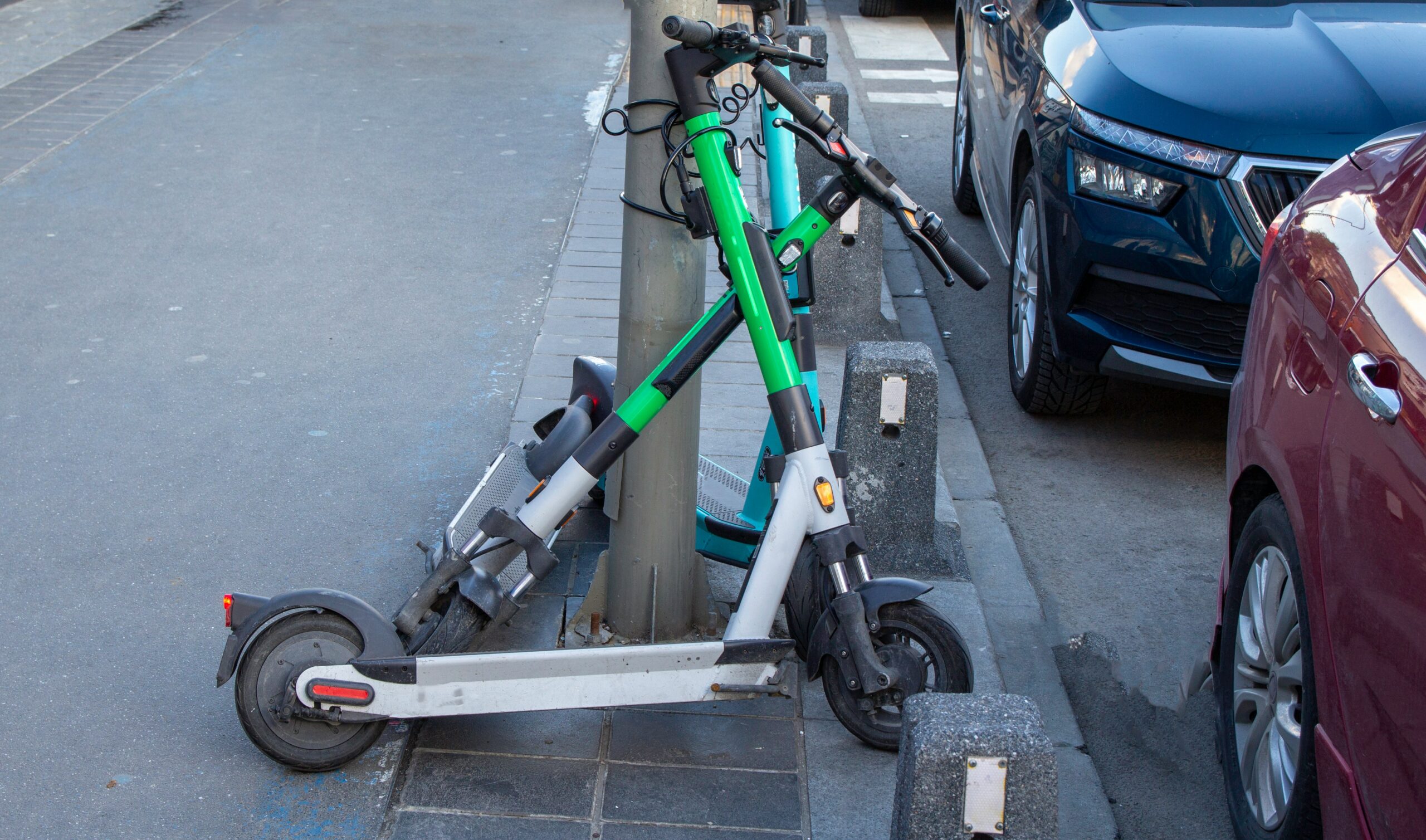 Two green and white electric scooters parked near a sidewalk, leaning against a post, with cars parked alongside them.