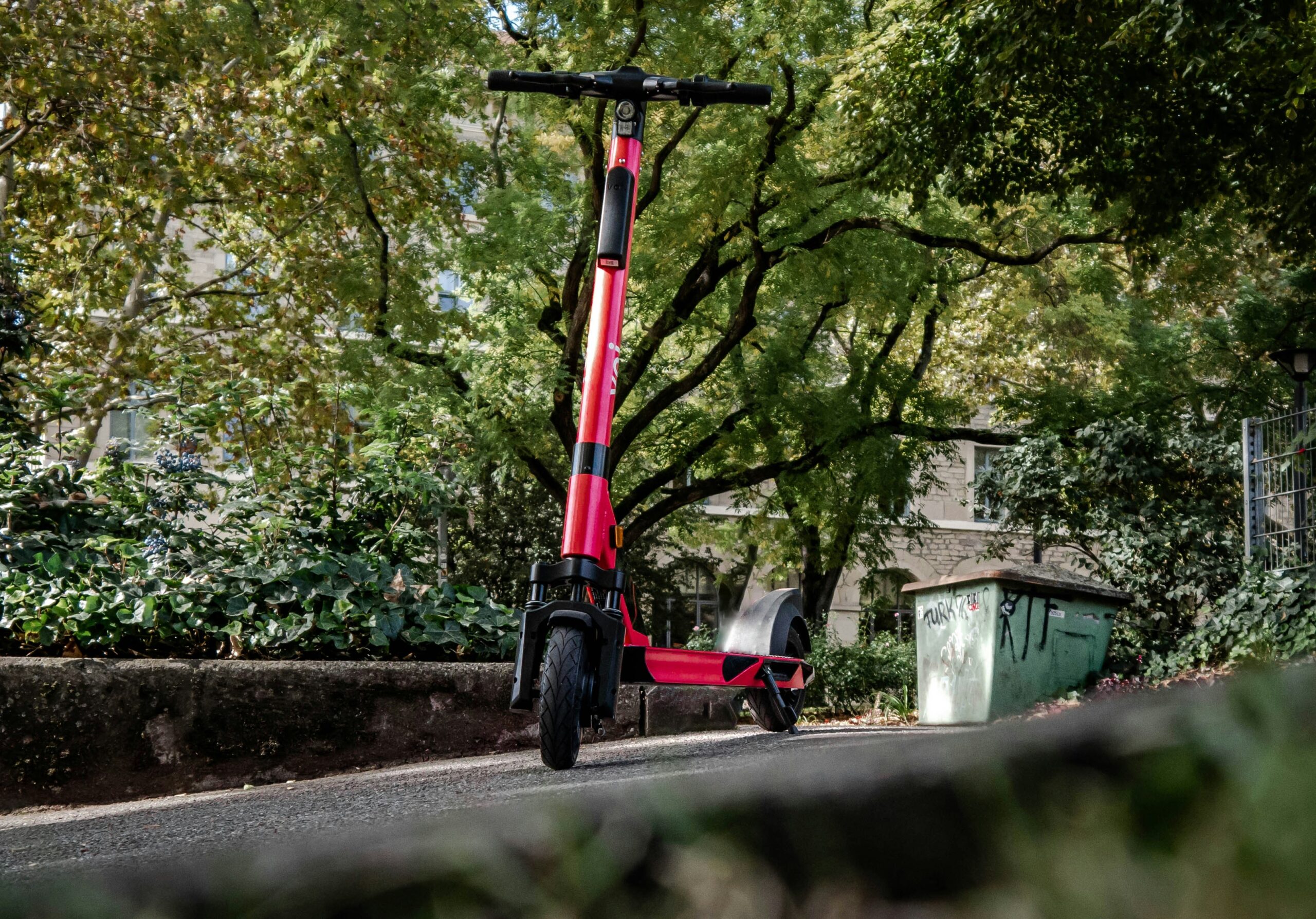 A red electric scooter parked on a quiet street surrounded by greenery and trees, with a trash bin in the background.