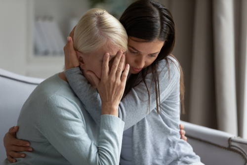 a young woman embracing an older woman who appears to be distressed. The older woman has her face in her hands, and the younger woman is leaning in closely, offering comfort and support. Both women are sitting on a couch, with a soft, empathetic mood conveyed through their body language. The scene suggests an emotional moment of grief, loss, or hardship, with the younger woman providing solace during a difficult time.