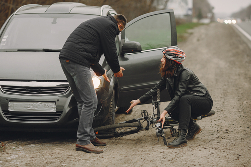 A cyclist sitting on the ground next to a fallen bicycle while a concerned driver kneels nearby next to an open car door after a roadside accident.