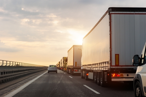 The trucks are closely aligned in a single lane, and the soft, golden light of the setting sun illuminates the scene. The sky is partially cloudy, casting a warm glow over the highway.