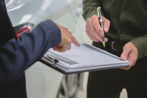 Two individuals reviewing and signing documents on a clipboard, likely related to a car accident or insurance claim, with a vehicle in the background.