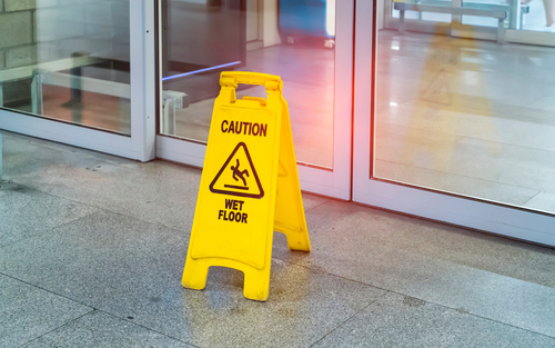 Yellow caution sign reading “Wet Floor” placed near a glass entrance.