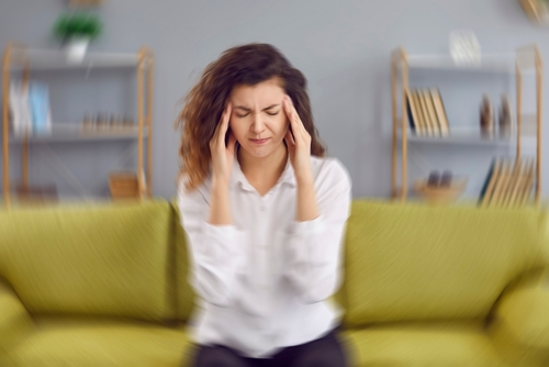 A woman sitting on a couch holding her head, appearing dizzy and in pain, showing symptoms of a brain injury.