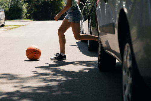 A child chasing a basketball across a residential street near parked cars, illustrating a potential pedestrian accident risk.