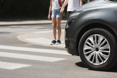 A car approaches a pedestrian crosswalk where two people stand at the edge of the road waiting to cross.