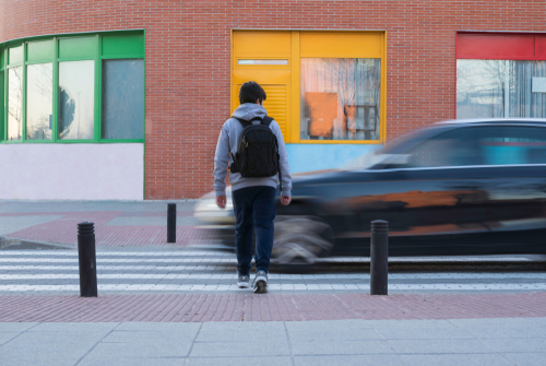 A student with a backpack waits at a crosswalk as a car speeds past on a city street near a brick building with colorful window frames.