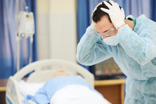 Distressed medical professional holding head in hands near a patient’s hospital bed.