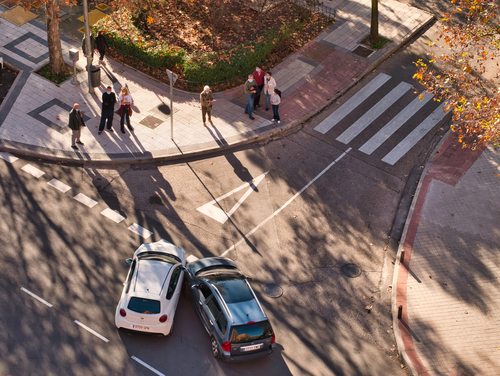 Aerial view of two cars colliding at an intersection near a crosswalk with pedestrians nearby.