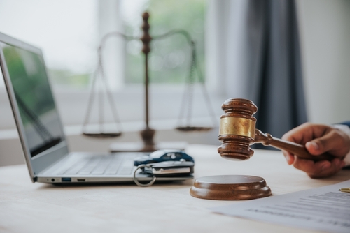 Close-up of a judge holding a wooden gavel beside a laptop and legal documents on a desk.