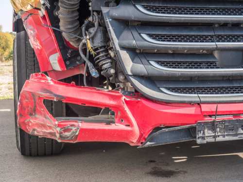 Close-up of a red semi-truck’s damaged front bumper and headlight area after a collision, revealing dents and broken components.
