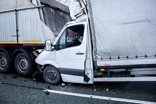 A white delivery truck crashed into the back of a large semi-trailer on a highway, showing severe front-end damage and debris scattered on the road.
