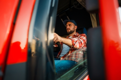 Male truck driver wearing a cap and plaid shirt sitting behind the wheel of a red truck, focused and looking ahead through the windshield.