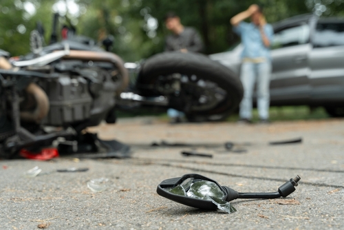 Close-up of motorcycle debris on the road after an accident, with blurred people and vehicles in the background.
