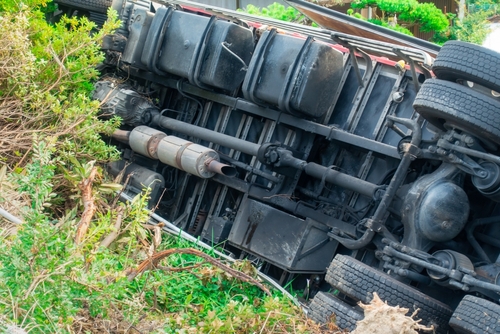 An overturned semi-truck lying on its side off the road, surrounded by damaged greenery after a serious crash.