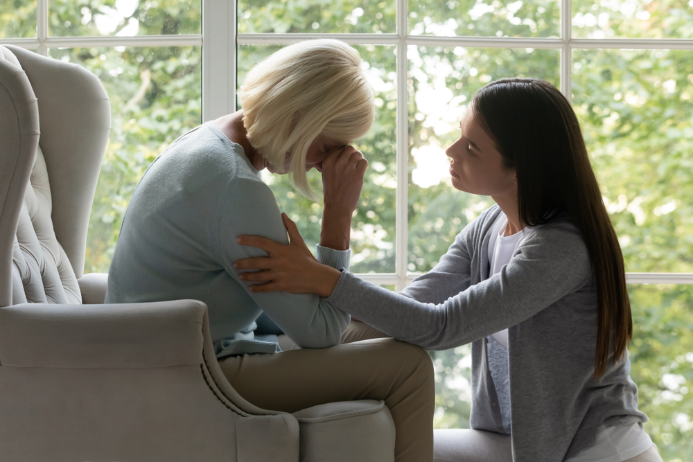 Empathic millennial lady soothing desperate crying mature mother, sitting in armchair. Side view upset grown up daughter comforting, supporting caring unhappy middle aged mommy felling unwell at home.