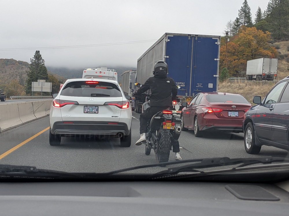 Grants Pass, OR – October 15, 2025 Traffic at a complete stop on a two-lane Oregon highway. A motorcycle ahead hesitates to lane split as fog and cloudy skies add tension to the quiet roadside scene.
